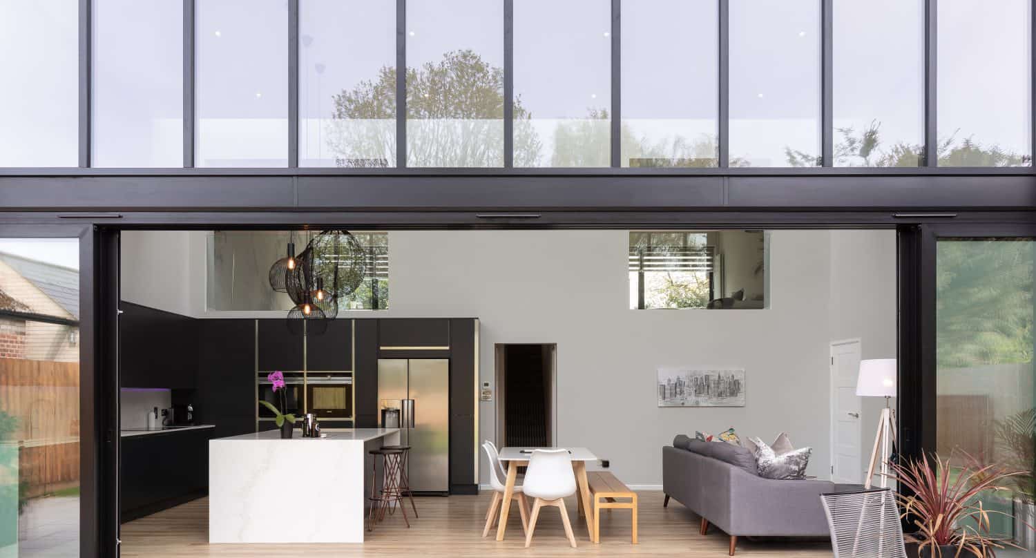 Modern open-plan living space with large bifold doors, featuring a kitchen with an island, a dining area, and a living area with a grey sofa. Outdoor seating and plants are visible on the patio.