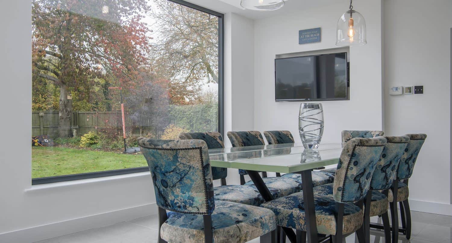 Modern dining room with a glass table, eight blue-patterned chairs, and large sliding doors overlooking a garden. Pendant lights and a wall-mounted TV add style, while natural light fills the space, creating a bright atmosphere.
