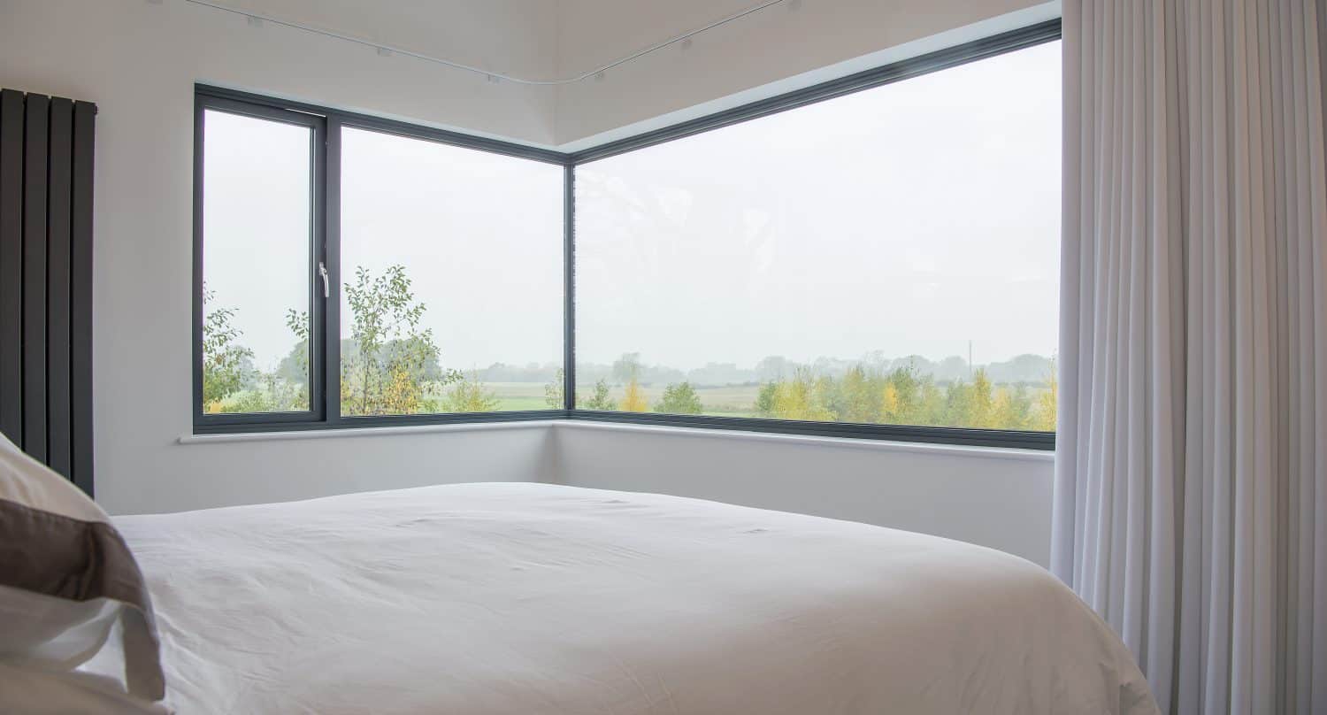 A modern bedroom with a large bed covered in white bedding features bespoke glass solutions in the form of floor-to-ceiling corner windows, offering views of a foggy landscape with trees and fields outside. White curtains partially frame the window.