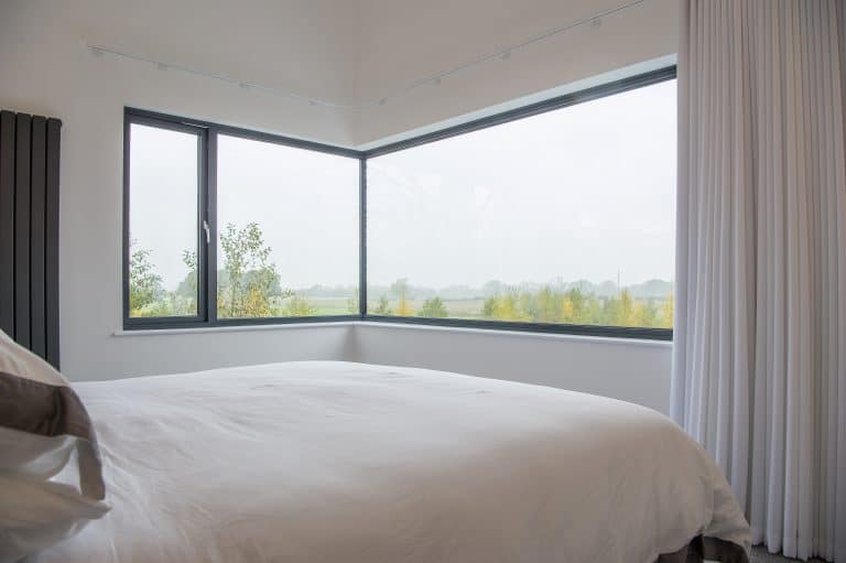 A modern bedroom with a large bed covered in white bedding features bespoke glass solutions in the form of floor-to-ceiling corner windows, offering views of a foggy landscape with trees and fields outside. White curtains partially frame the window.
