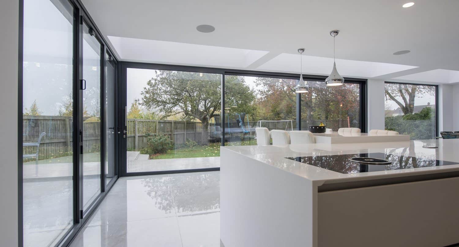 A modern kitchen with a large white island and breakfast bar, featuring floor-to-ceiling bifold doors that open to a garden with trees and a fence, letting in abundant natural light.