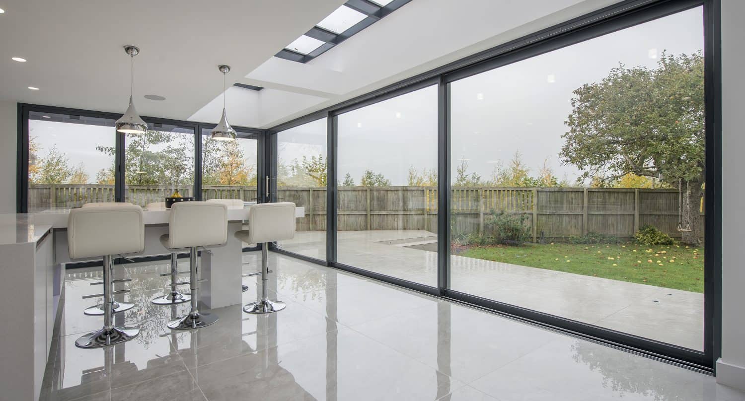 Modern kitchen with white bar stools, pendant lights, and large floor-to-ceiling sliding doors opening to a fenced backyard with trees and a lawn. The room features glossy tiled floors, skylights above, and bespoke glass solutions throughout.