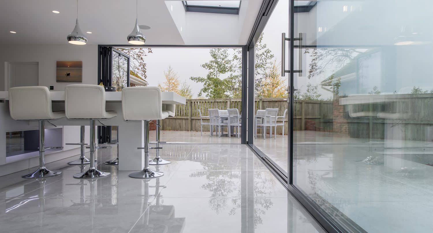 Modern kitchen with white bar stools and pendant lights, featuring bespoke glass solutions including large sliding doors that open to a patio with an outdoor dining table, all surrounded by a wooden fence and trees.