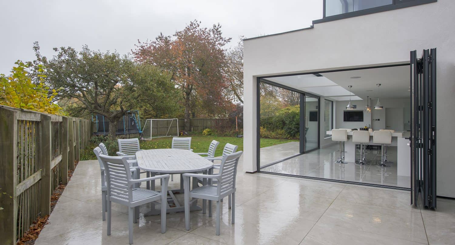 A modern house with large bifold doors opens to a patio featuring a round table and six chairs. The patio overlooks a garden with trees and a soccer goal, and the ground appears wet from recent rain.
