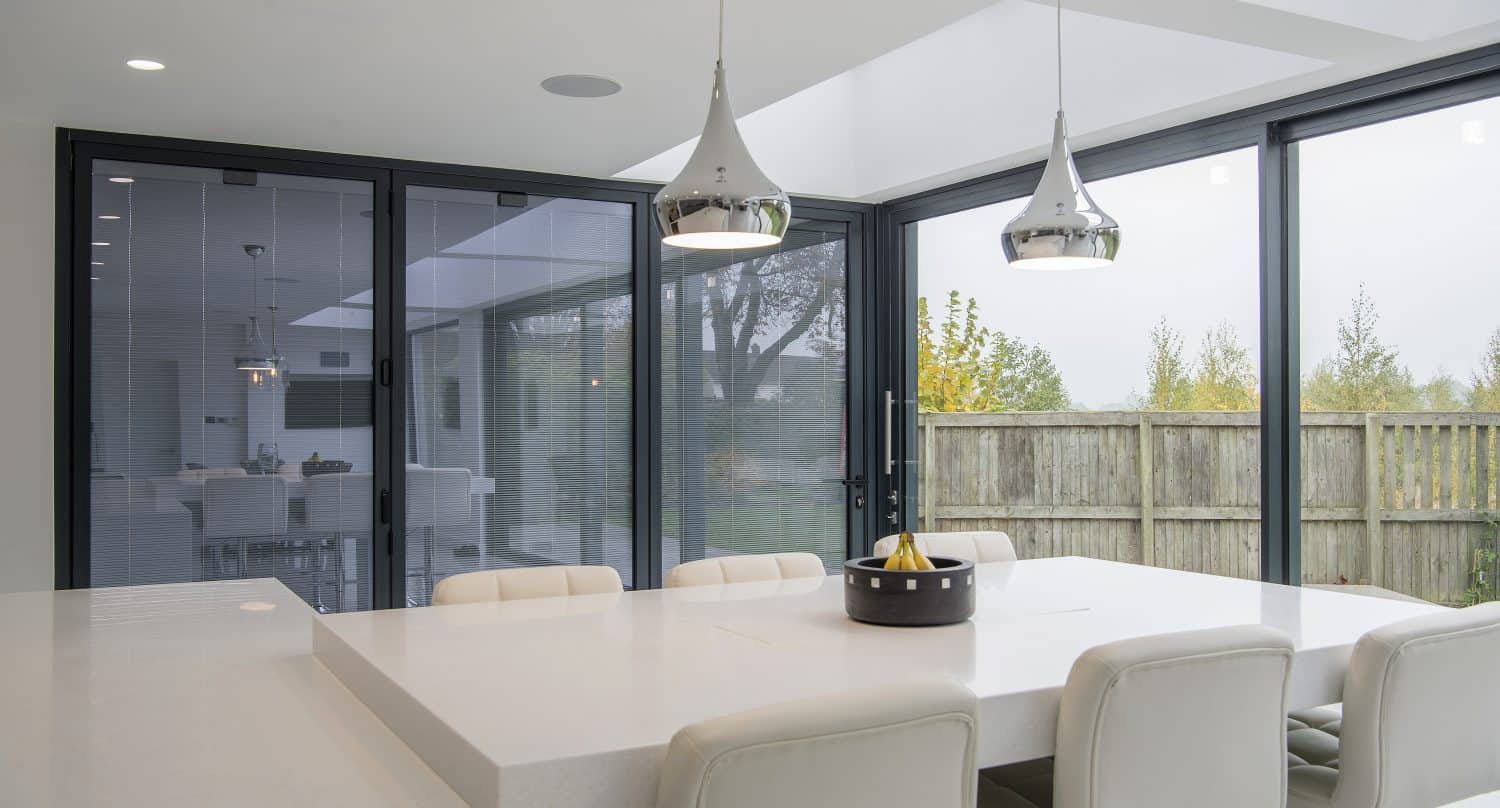 Modern dining area with a white table and cushioned chairs, pendant lights above, and large bifold doors offering a view of a wooden fence and trees outside. A fruit bowl sits on the table.