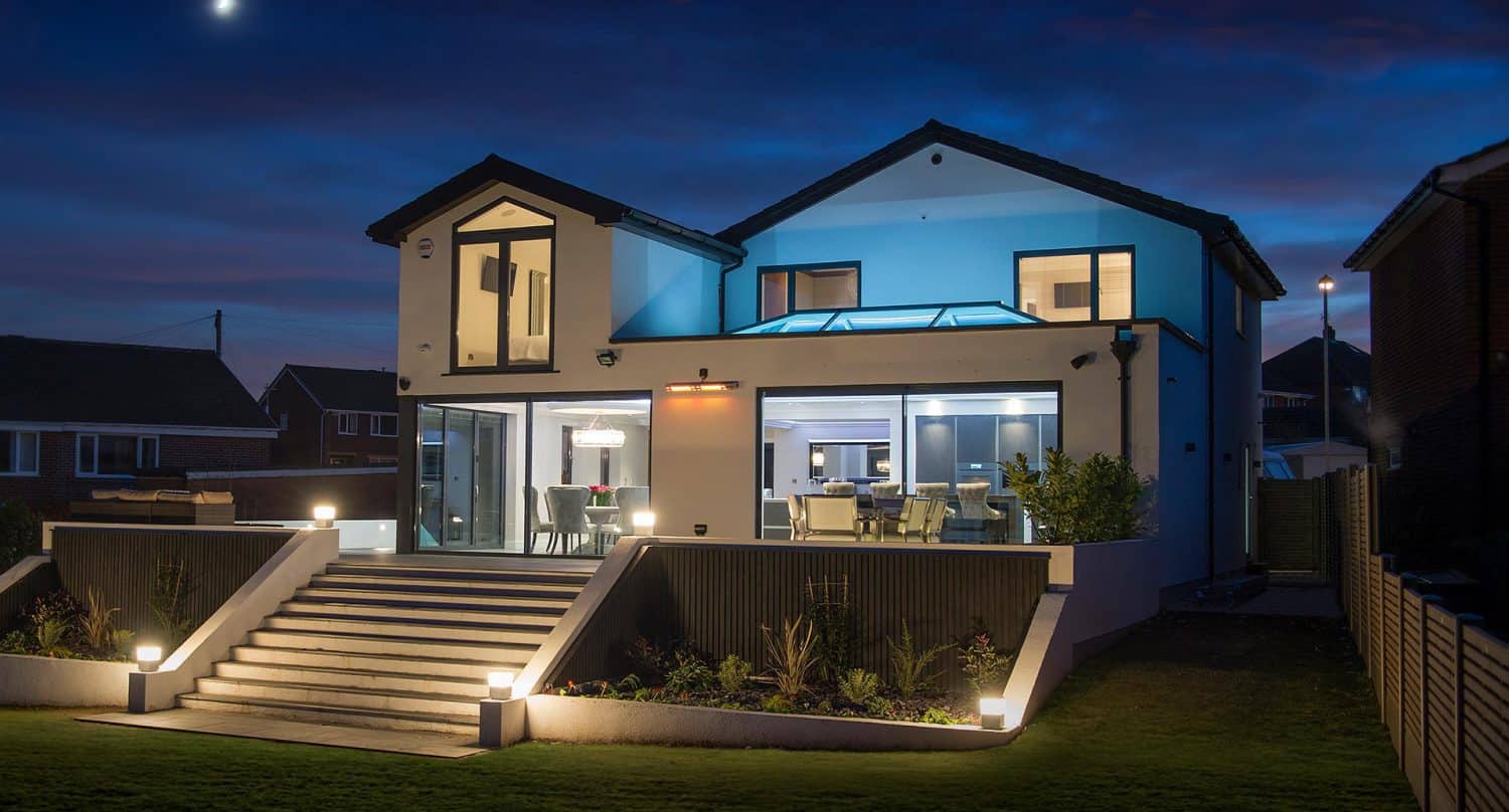 Modern two-story house with large windows and outdoor lighting, illuminated at night. Wide steps lead to stylish front doors, while interior lights reveal a contemporary dining and living area. A crescent moon graces the sky above.