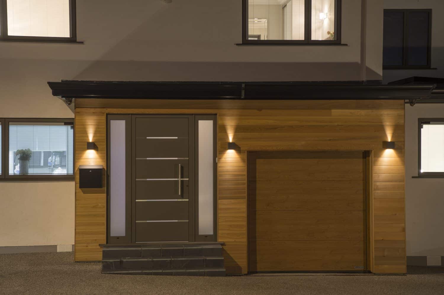 Modern house entrance at night with a wooden door, matching wood-clad walls, a mailbox, and bespoke glass solutions illuminating the entryway. Adjacent garage door also features a wooden finish.