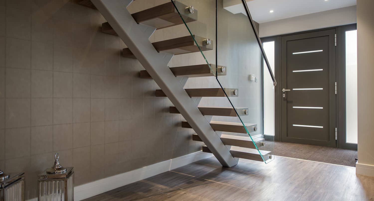 Modern hallway with wooden flooring, a floating wooden staircase with glass railing, taupe walls, decorative lanterns, and a dark front door with horizontal glass panels. Natural light enters from windows beside the door and sleek sliding doors.