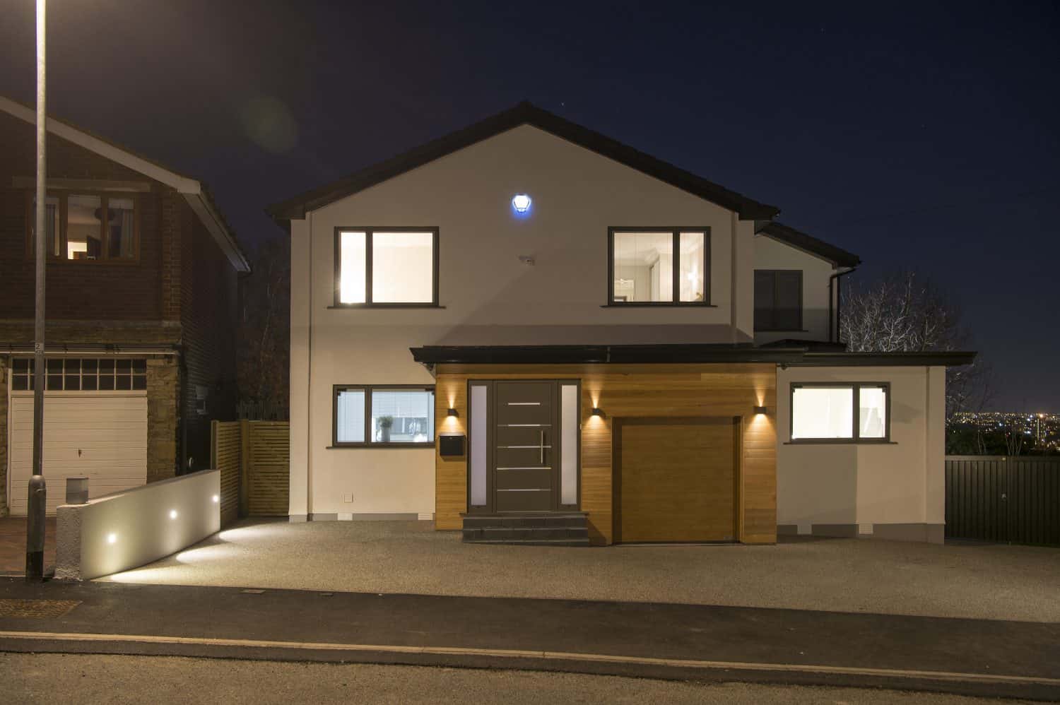 A modern two-story house with large windows and a lit entrance, photographed at night. The exterior features white walls, wooden accents, and striking front doors, with a driveway and a garage to the right.