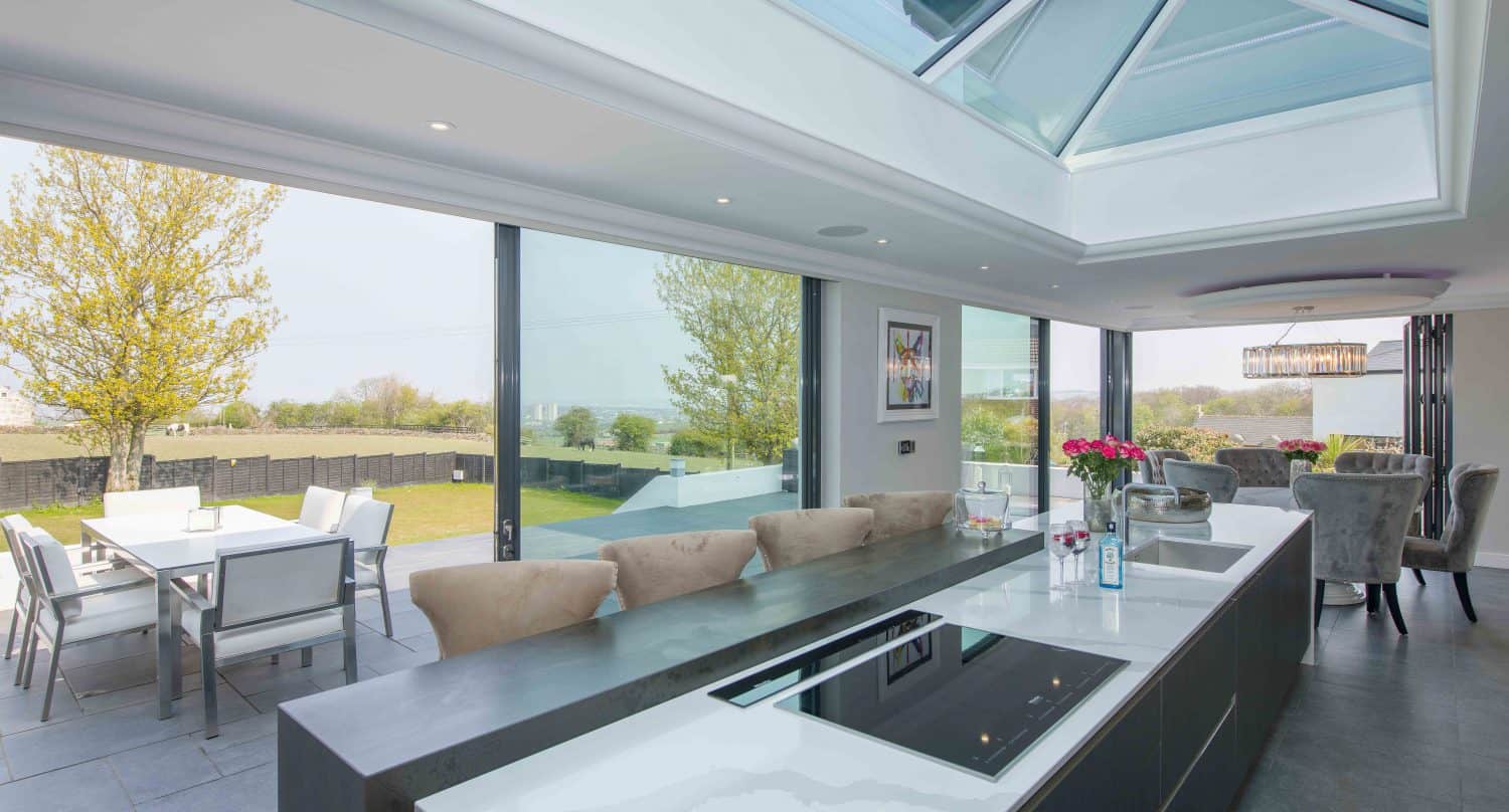 Modern kitchen with a large island, built-in stovetop, and sink. Bifold doors open to a patio with outdoor dining, letting in natural light. Skylights and trees are visible outside. Roses sit on the counter.