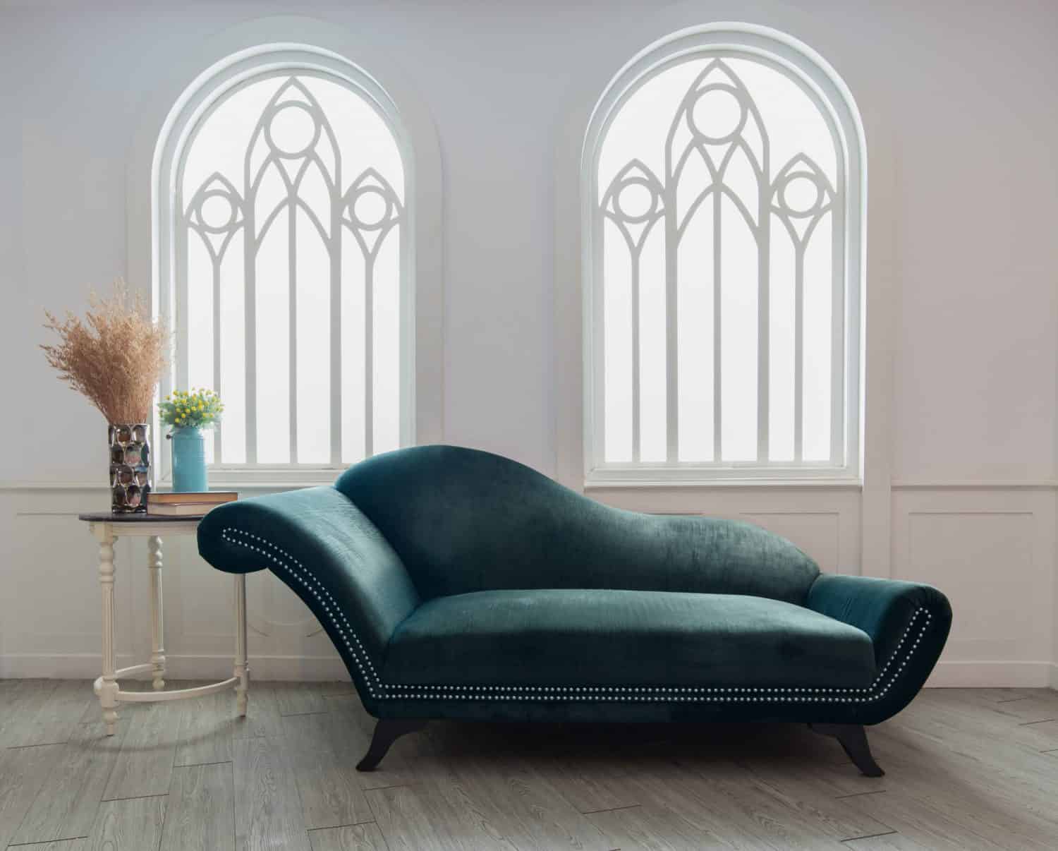 A dark teal chaise lounge with silver studs sits on a light wood floor between two arched windows and bespoke glass solutions bifold doors. A small round table holds a vase of dry grass and a bouquet of yellow flowers.