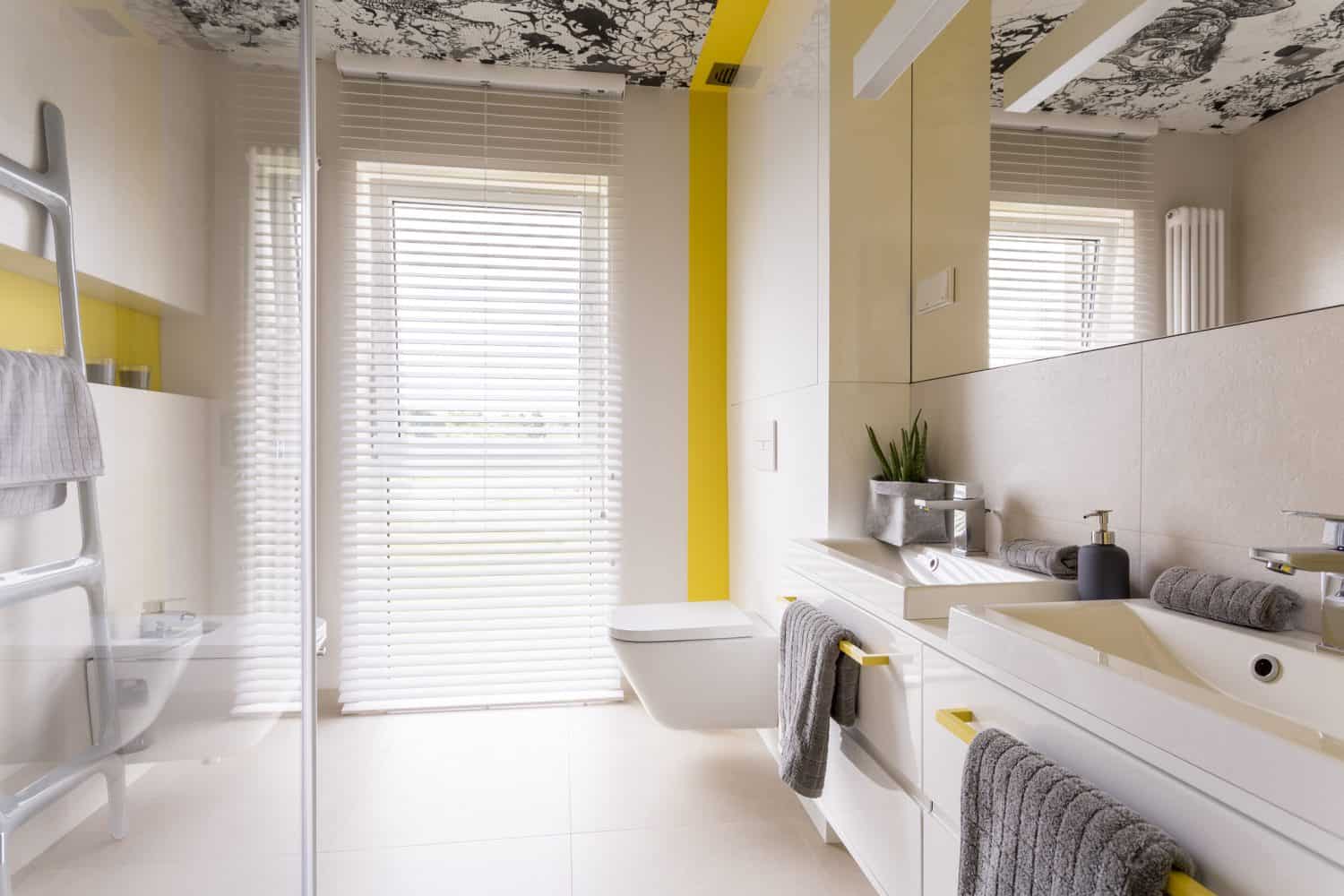 Fresh and bright contemporary bedroom with beige tiles, mirror, graphic ceiling, double sink, toilet, yellow decor and window blinds
