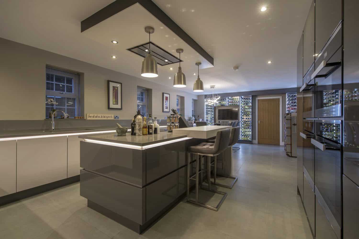 Modern kitchen with glossy grey cabinets, a large island with bar stools, pendant lighting, built-in appliances, and a wine display wall in the background. Bespoke glass solutions enhance the sleek, well-lit spaces contemporary design.