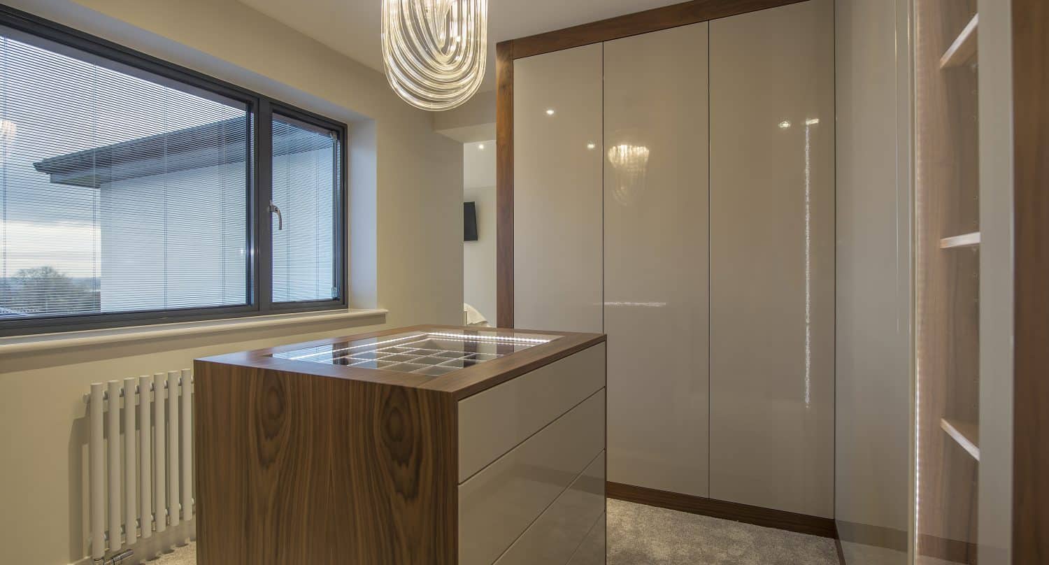 A modern walk-in closet with glossy beige wardrobes, a wooden island with drawers, carpeted floor, sliding doors, a large window with blinds, and a chandelier overhead.