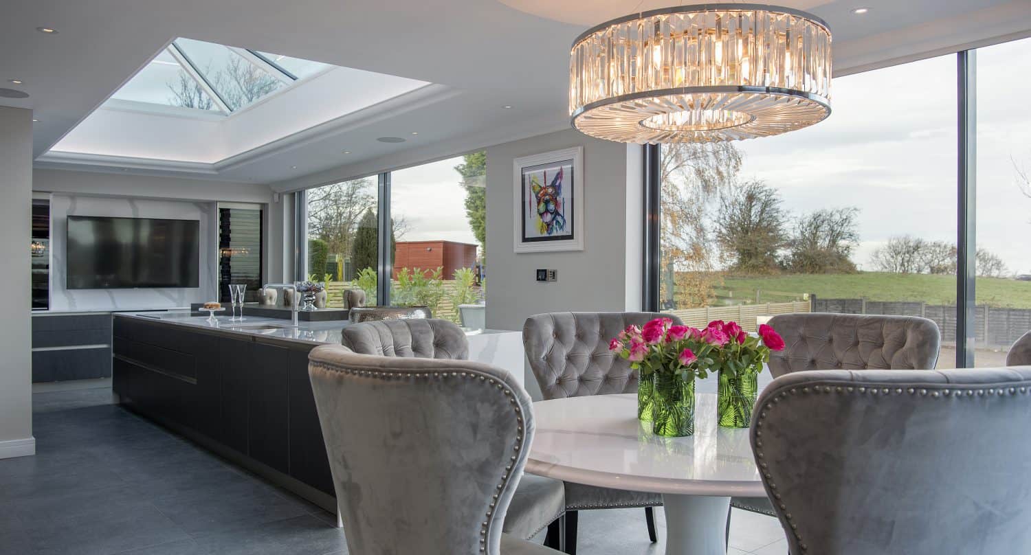 Modern dining area with a round white table, gray velvet chairs, and pink flowers in vases. Large windows, skylights, and stylish sliding doors let in natural light. A chandelier hangs above the table; a sleek kitchen is visible in the background.