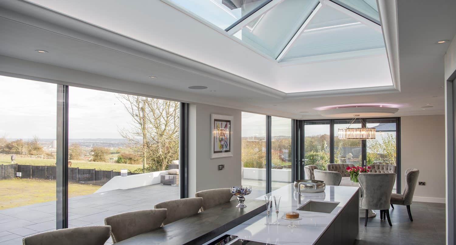 Modern kitchen and dining area with a large skylight, marble island, elegant chairs, and bifold doors offering a scenic view of the outdoors. Natural light fills the spacious, contemporary room.