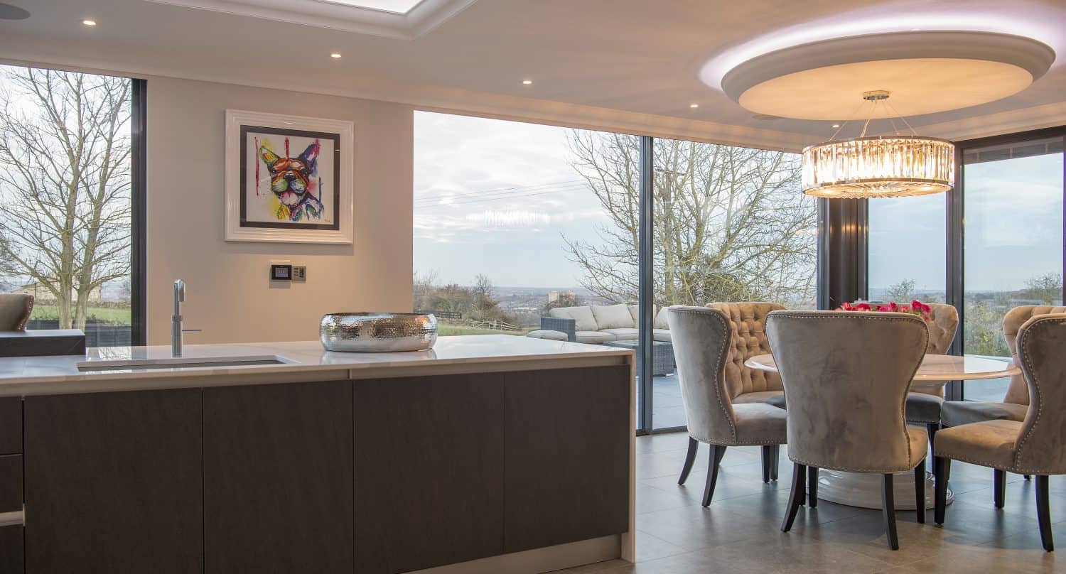 Modern open-plan kitchen and dining area with large windows, sliding doors, a circular dining table with plush chairs, contemporary chandelier, wall art, and views of trees and a patio outside.