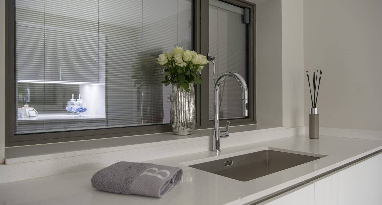 A modern kitchen countertop with a built-in sink, a silver faucet, a vase of white roses, a gray towel, and a reed diffuser. Large sliding doors and a window with blinds bring in natural light above the counter.