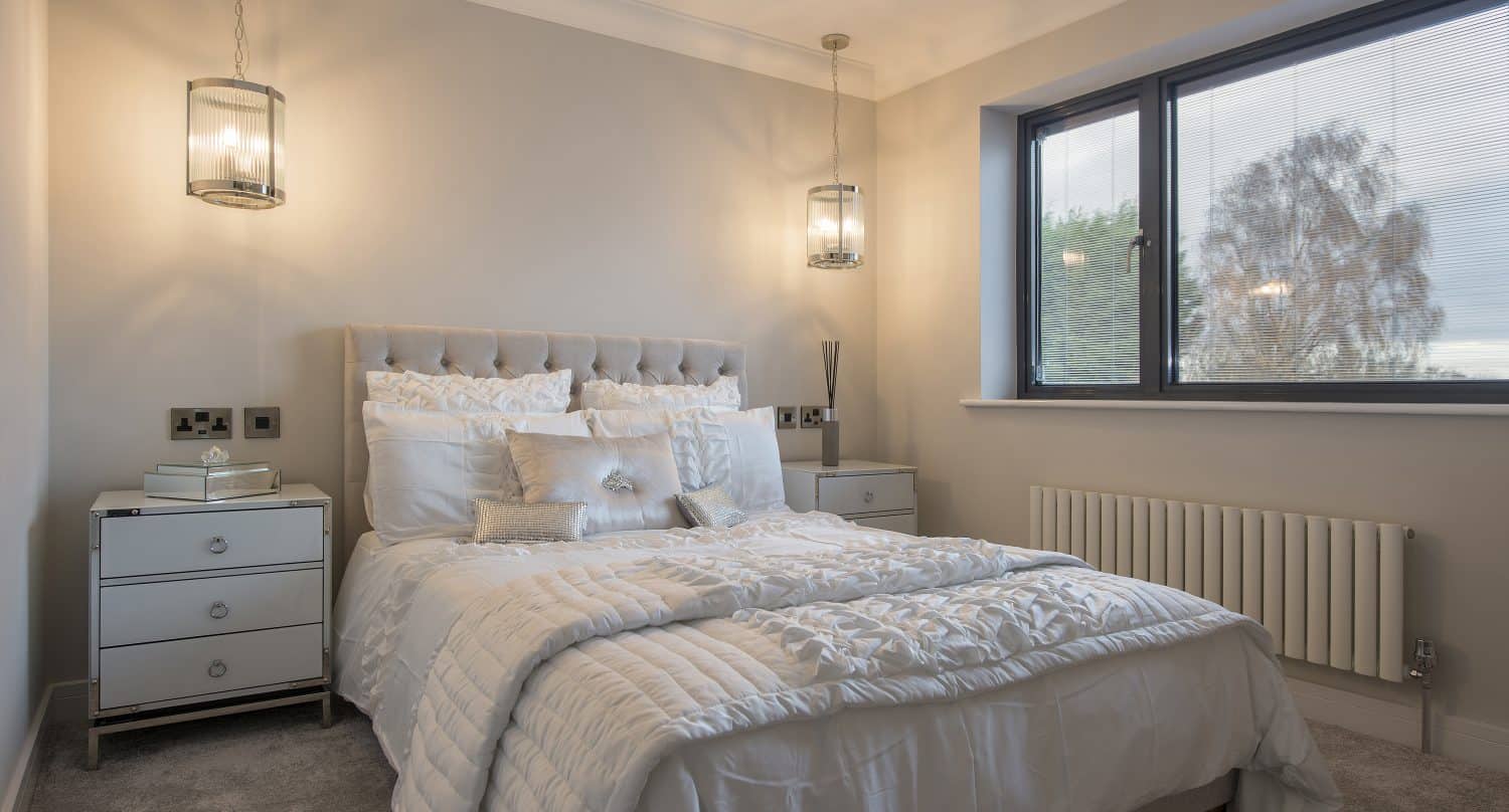 Modern bedroom with a double bed dressed in white bedding, two bedside tables with lamps, and a large window letting in natural light. Elegant sliding doors enhance the neutral decor and add to the room’s cozy, inviting atmosphere.