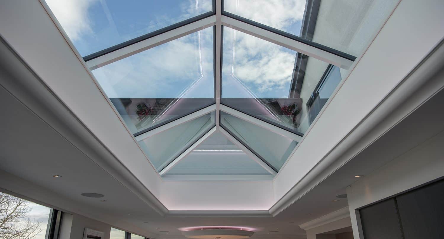 A modern interior with a large bespoke glass skylight ceiling lets in natural light and offers a view of a partly cloudy blue sky. The room features white walls, recessed lighting, and contemporary decor.
