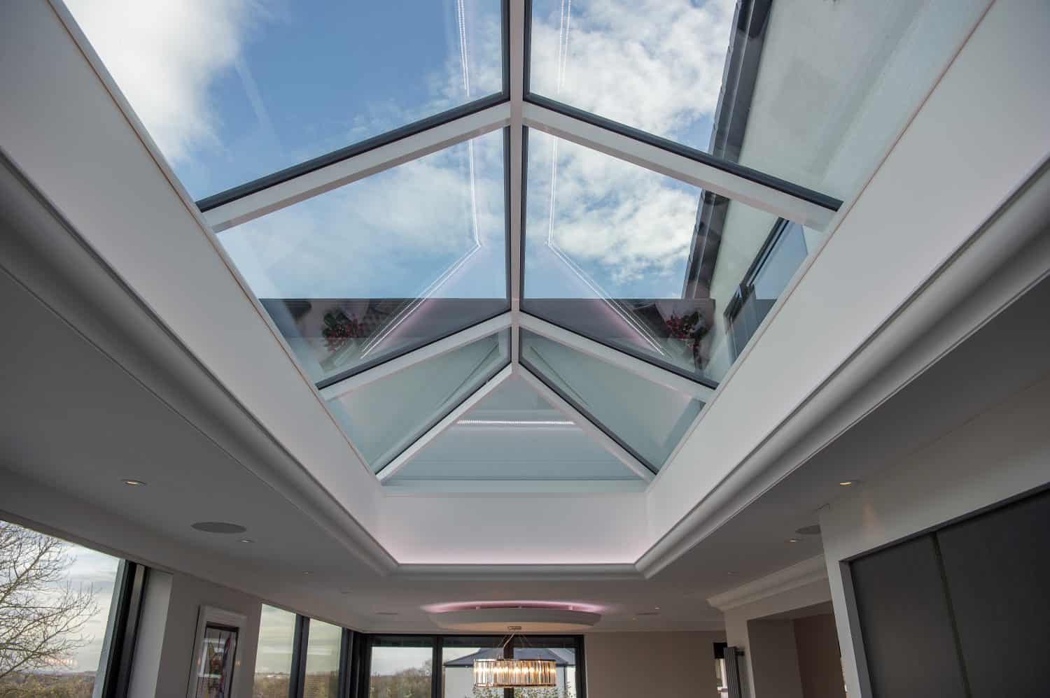 A modern interior with a large bespoke glass skylight ceiling lets in natural light and offers a view of a partly cloudy blue sky. The room features white walls, recessed lighting, and contemporary decor.