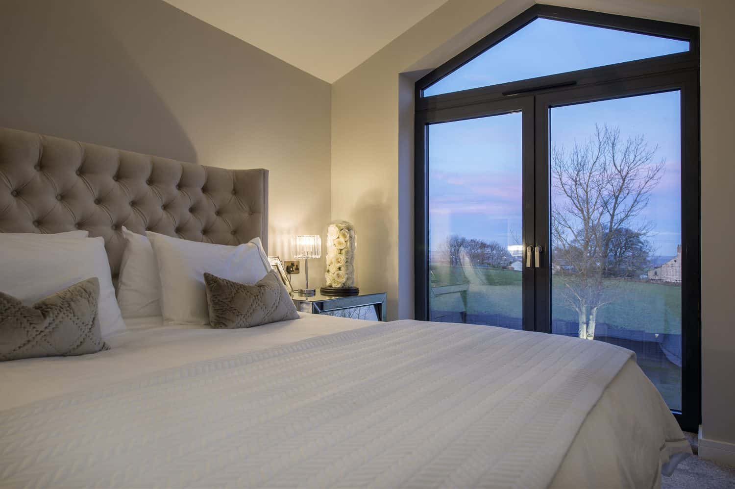 A cozy bedroom with a large bed, tufted headboard, and two decorative pillows sits beside a lamp-lit table. Bespoke glass solutions in the large doors reveal a tree and colorful evening sky just outside.