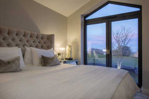 A cozy bedroom with a large bed, tufted headboard, and two decorative pillows sits beside a lamp-lit table. Bespoke glass solutions in the large doors reveal a tree and colorful evening sky just outside.