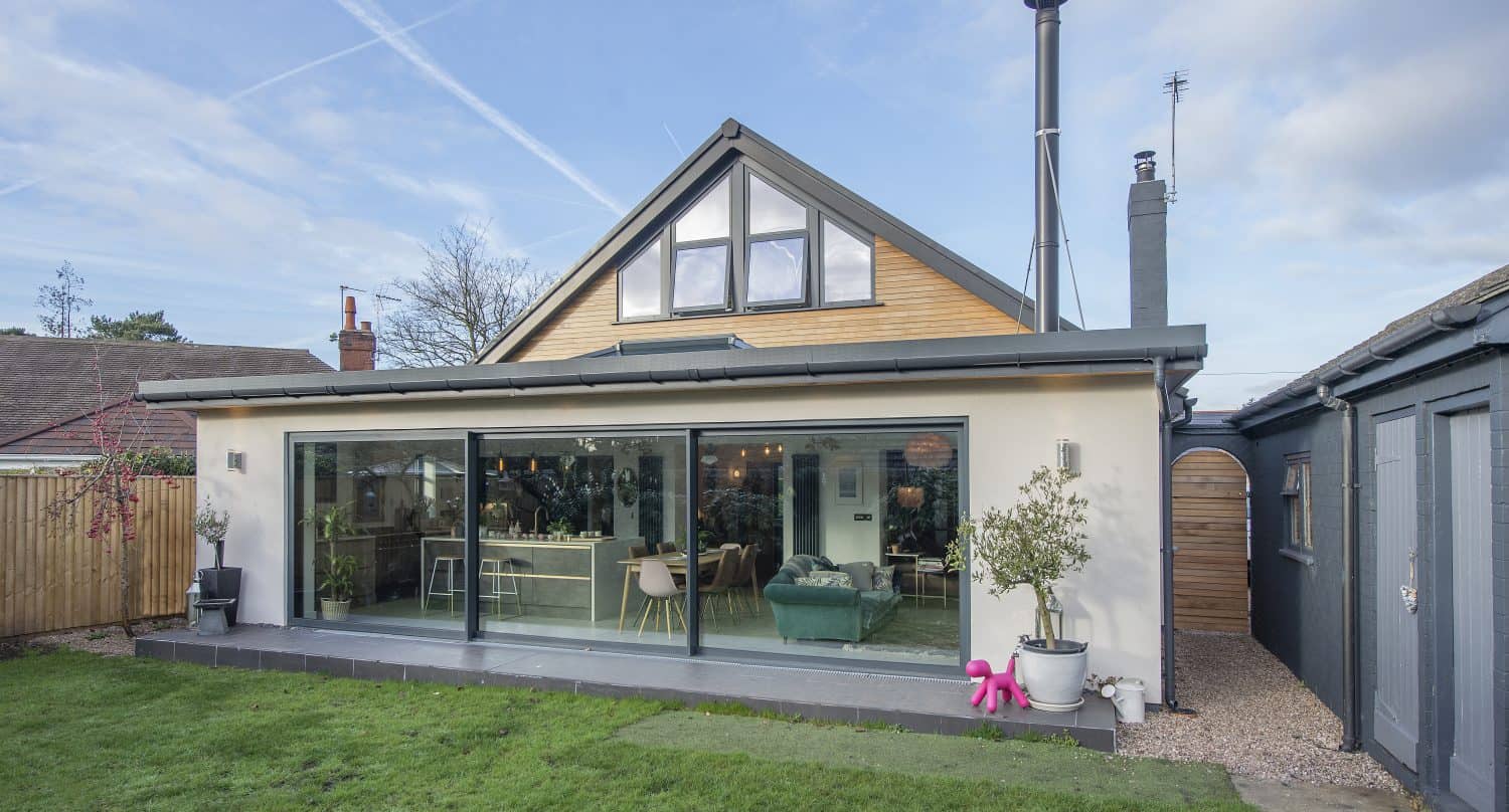 A modern house with large bifold doors opening onto a green lawn, featuring an upper loft with a big triangular window and a patio with potted plants. The sky is clear with some clouds and vapor trails.