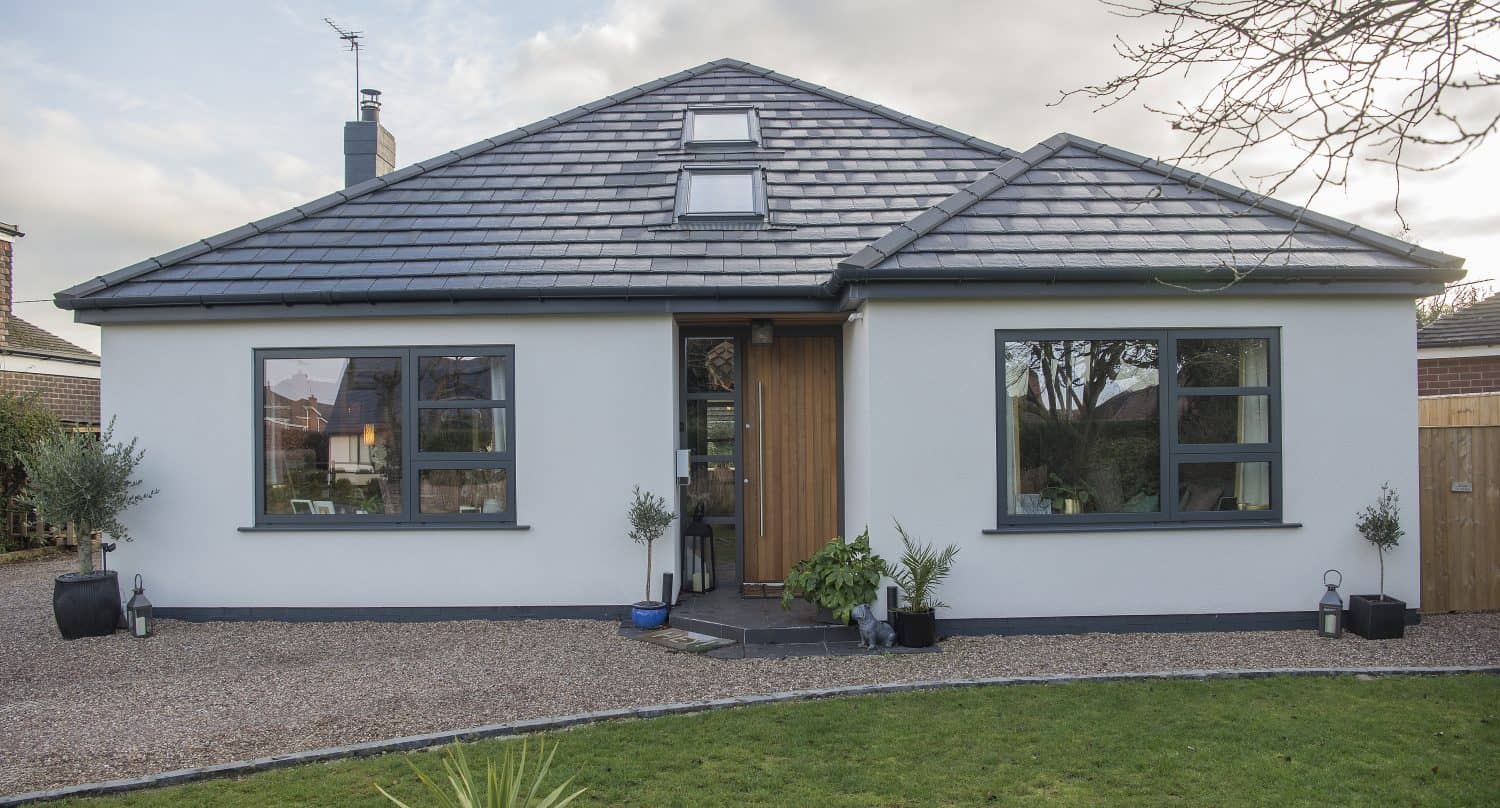 A modern single-story house with a gray tiled roof, white exterior walls, large windows, and sliding doors. The wooden front door opens to a gravel path, potted plants, and a green lawn.