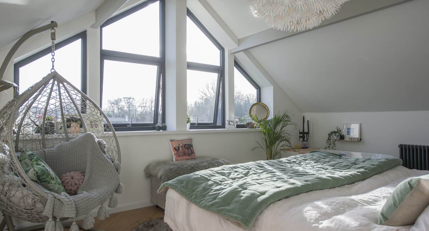 A cozy attic bedroom with large triangular windows, bespoke glass solutions, a hanging wicker chair with cushions, a fluffy rug, a green comforter on the bed, plants, and soft natural light streaming in.
