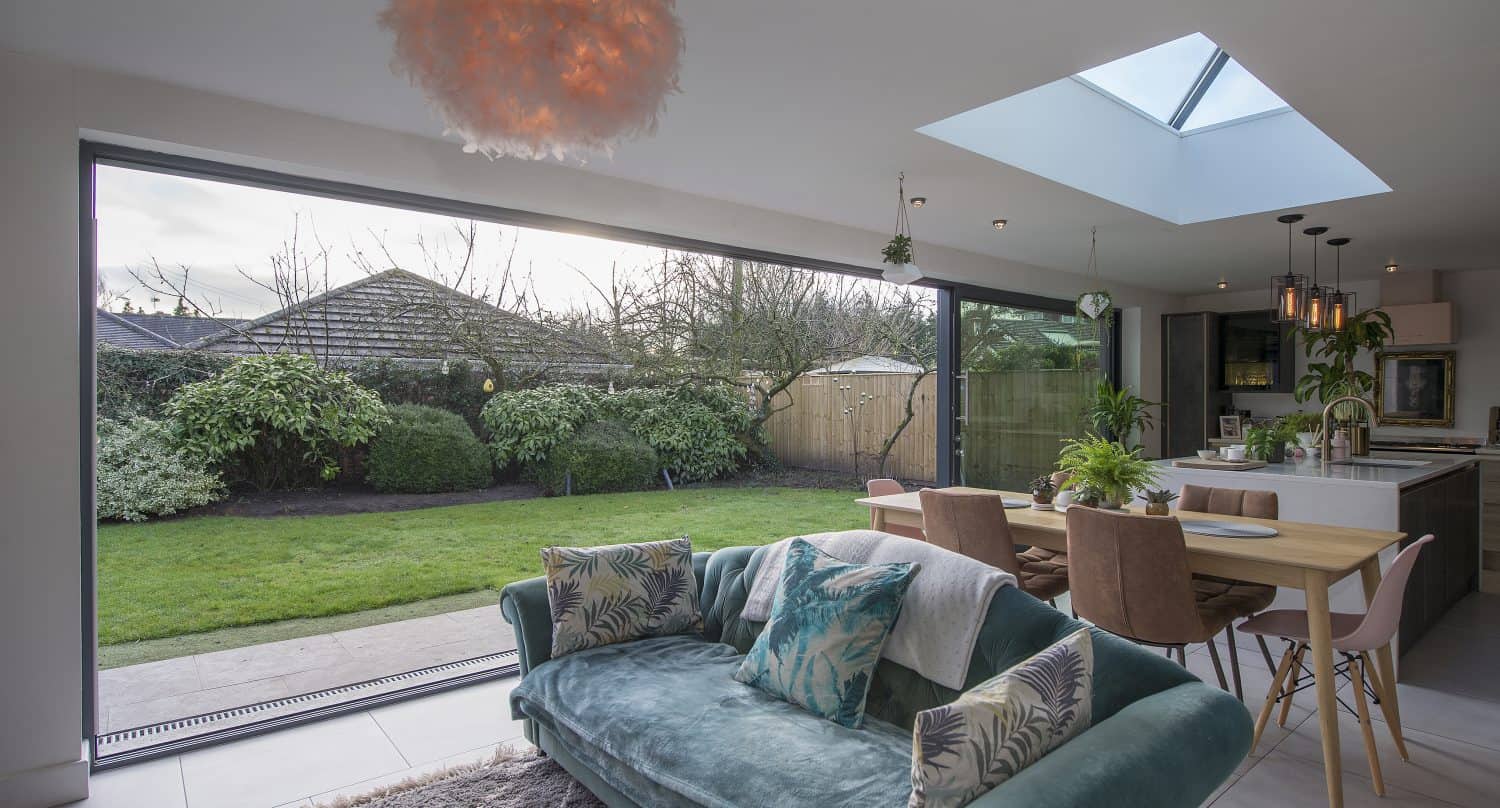 A modern, bright living and dining area with a green velvet sofa, a wooden table with pink chairs, a glass roofing skylight, and large sliding doors opening to a lush, green garden.