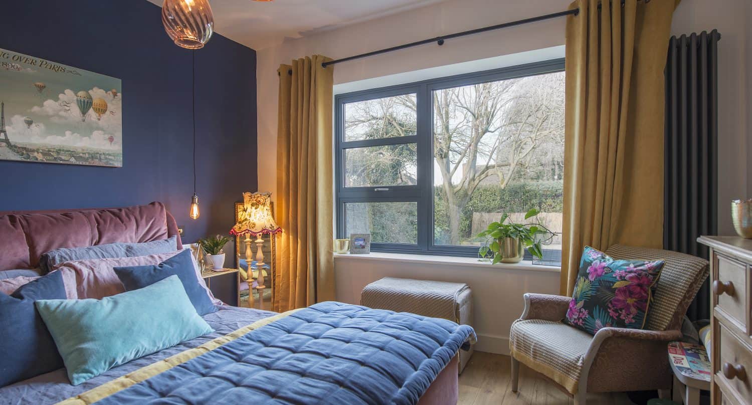 A cozy bedroom with a large window featuring bespoke glass solutions, blue and yellow bedding, pink headboard, blue accent wall, yellow curtains, decorative pillows, plants, and a patterned armchair beside a dresser. Warm pendant lights hang from the ceiling.