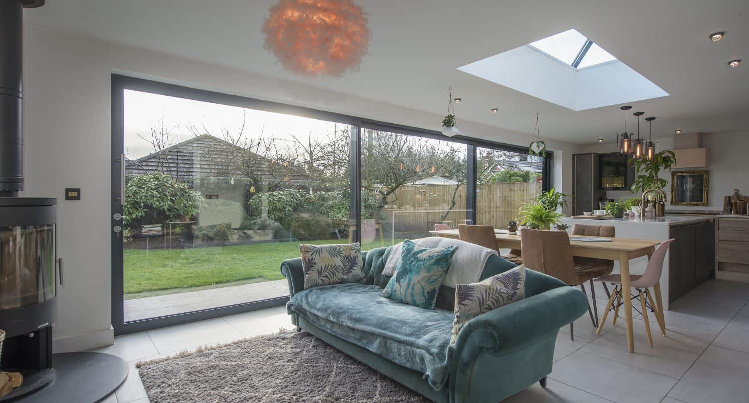 A modern open-plan living and dining area features a teal velvet sofa, a fluffy rug, bespoke glass solutions like bifold doors overlooking the garden, a skylight, dining table with pink chairs, and lush indoor plants.