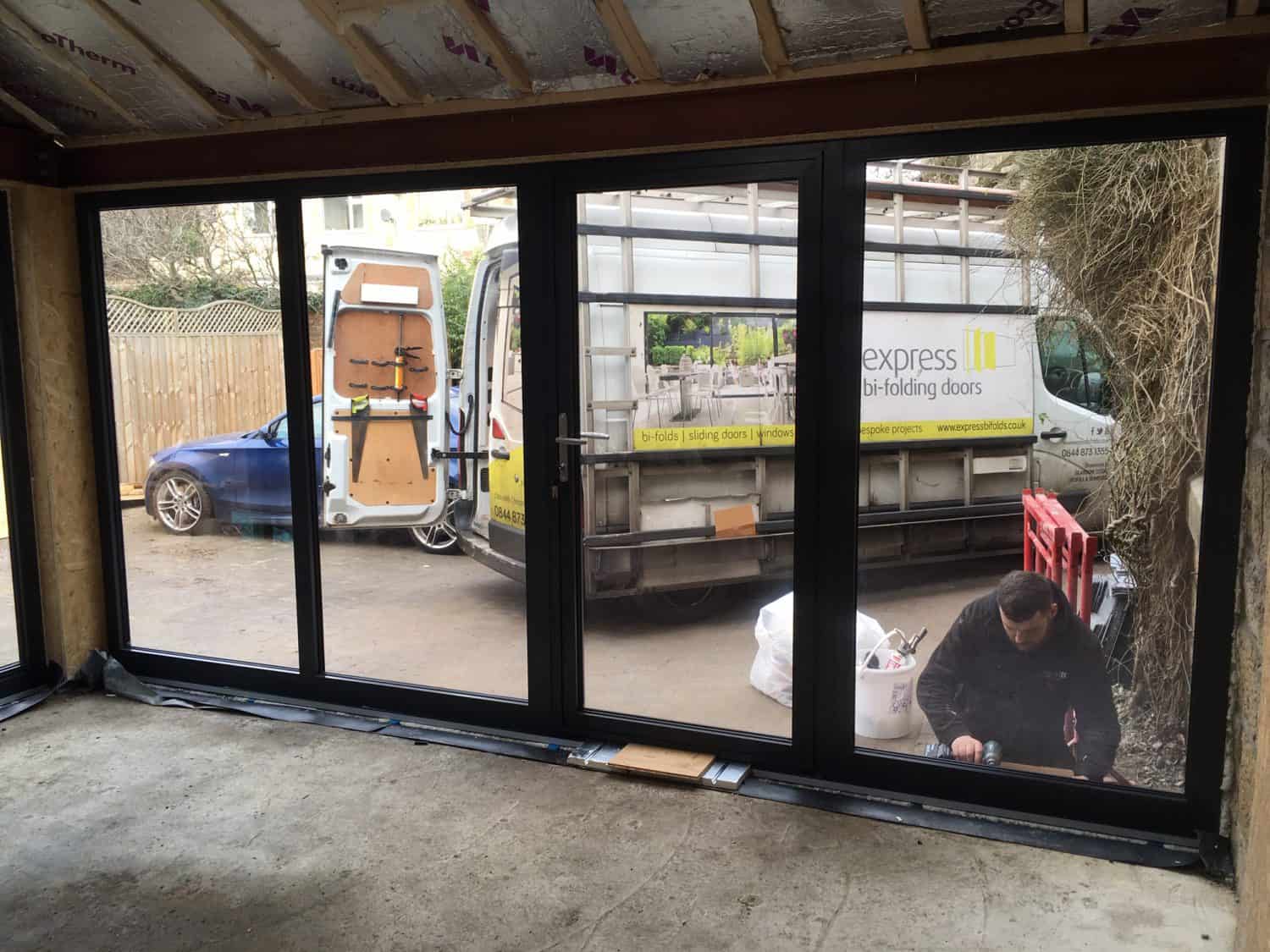 A person installs large black-framed glass folding doors in a house, creating bespoke glass solutions. Outside, a van with “Express Bi-Folding Doors” branding is parked, alongside a blue car and building materials.