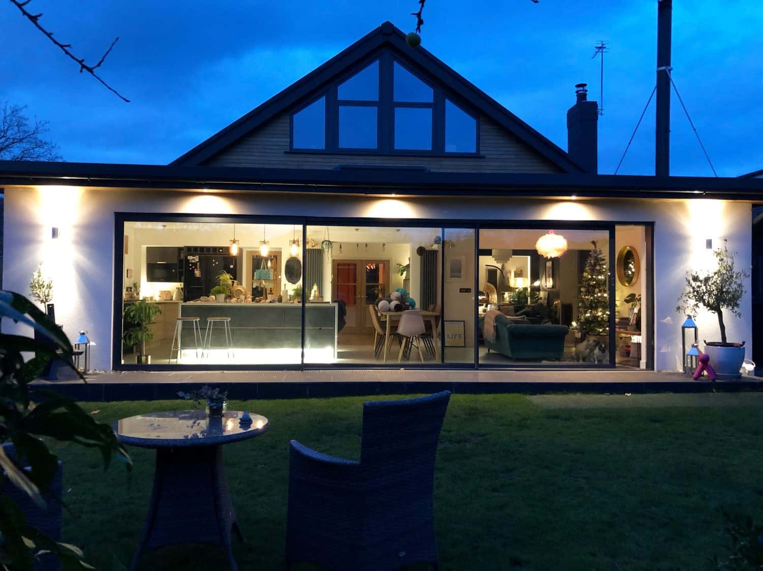 A modern house with large bifold doors reveals a warmly lit interior, including a kitchen, living area, and decorated Christmas tree, seen from a garden with outdoor seating at dusk.