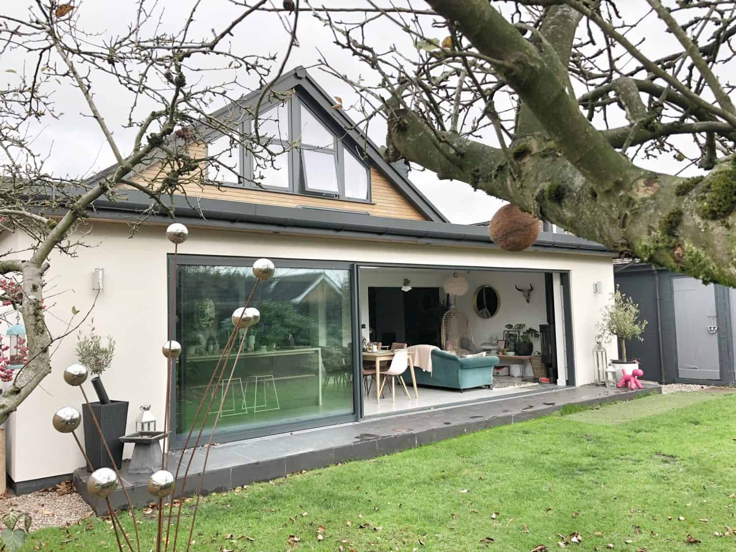 A modern house with large sliding doors opens onto a green lawn. A tree with bare branches and silver garden ornaments is in the foreground. Inside, a blue sofa and dining table are visible beneath elegant glass roofing.