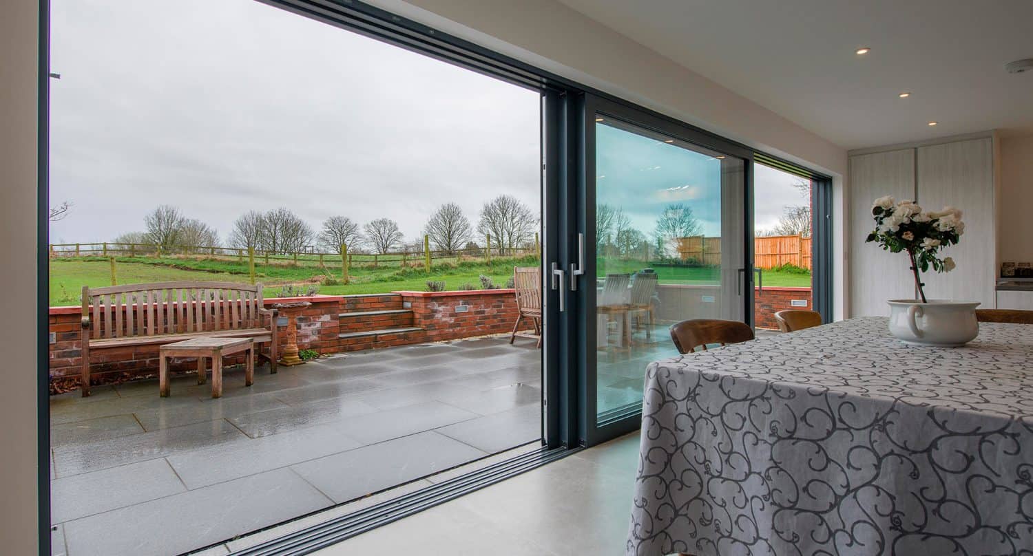 Modern dining area with a table covered by a patterned cloth, featuring large bifold doors that open to a patio with outdoor seating and views of a grassy yard and trees.