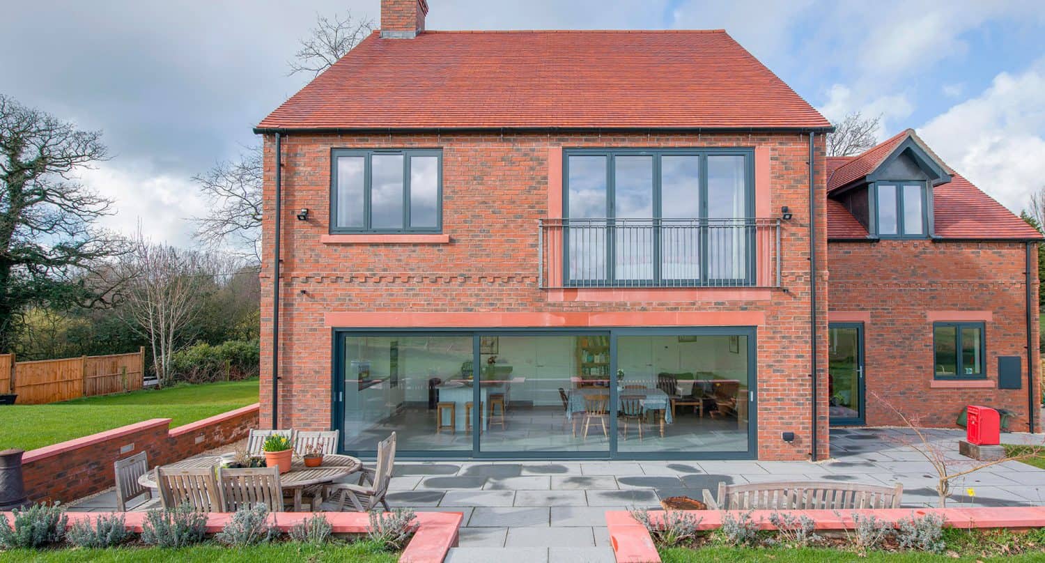 Modern two-story brick house with large bespoke glass solutions, including bifold doors and expansive windows, a red-tiled roof, and a spacious patio area with outdoor seating, surrounded by a well-maintained lawn and garden.