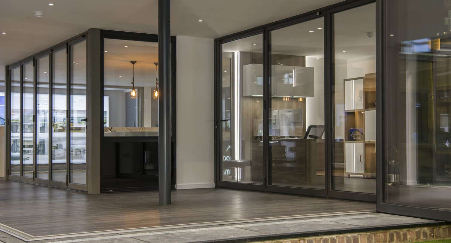 Modern interior with large bifold doors, wood flooring, and recessed lighting. The space features a visible kitchen area with white cabinets and pendant lights, creating an open and contemporary design.