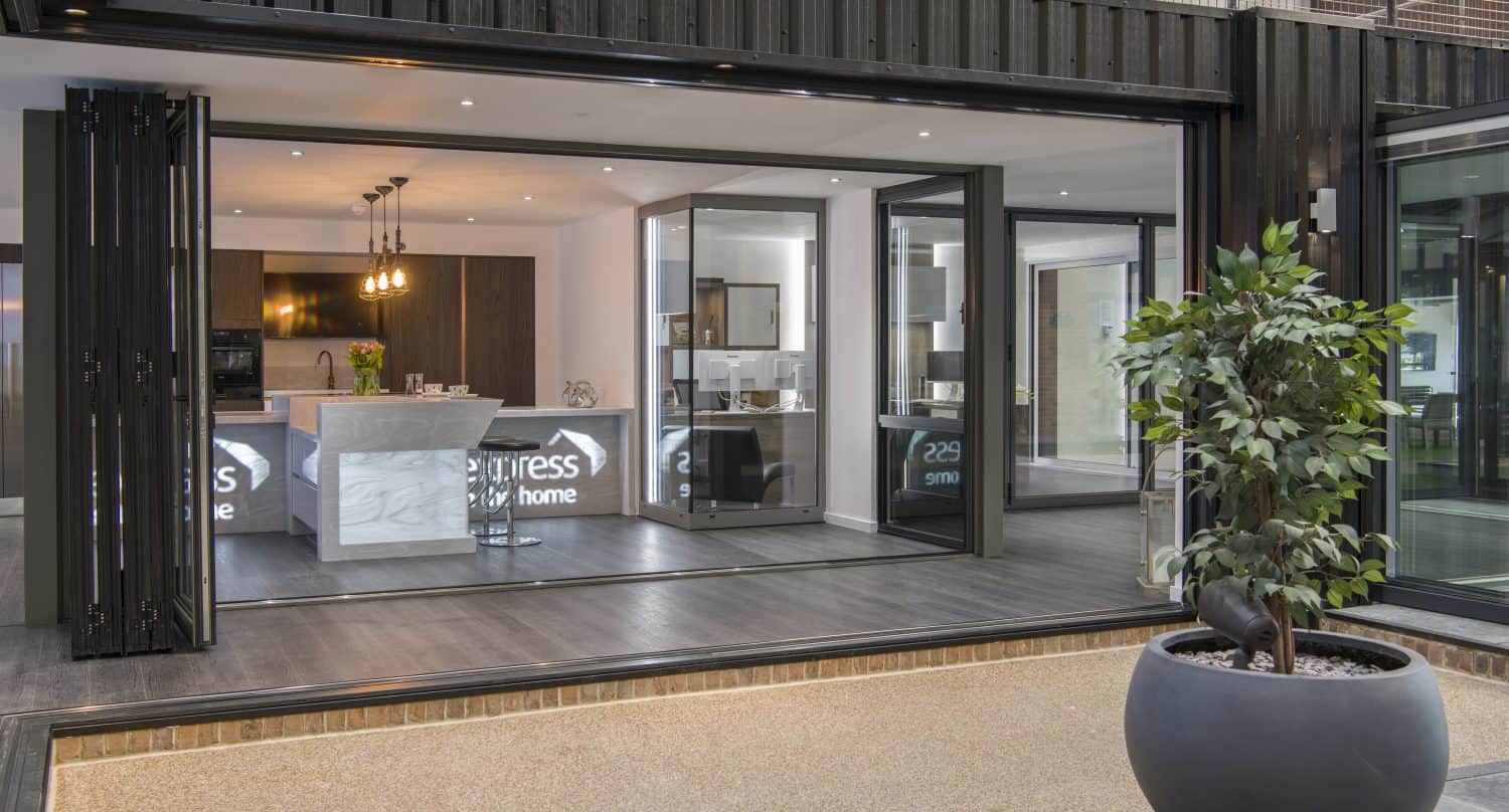 Modern open-concept kitchen and living area with large bifold doors opening to a patio; a potted plant sits in the foreground on a stone surface.