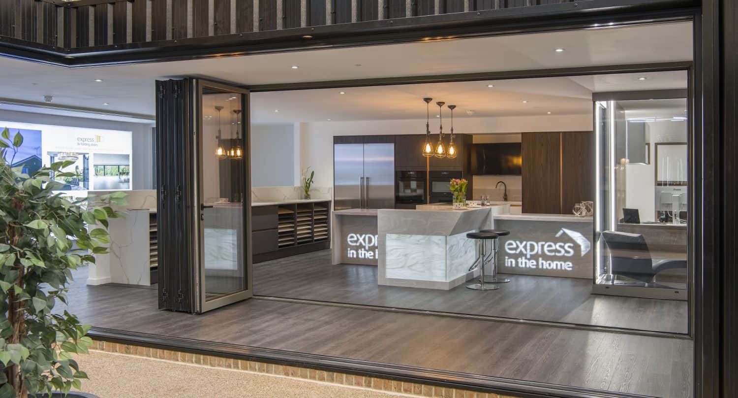 Modern kitchen showroom with large bifold doors open, revealing a stylish space with marble countertops, dark cabinets, pendant lights, and express in the home text on the island. An indoor plant is visible in the foreground.