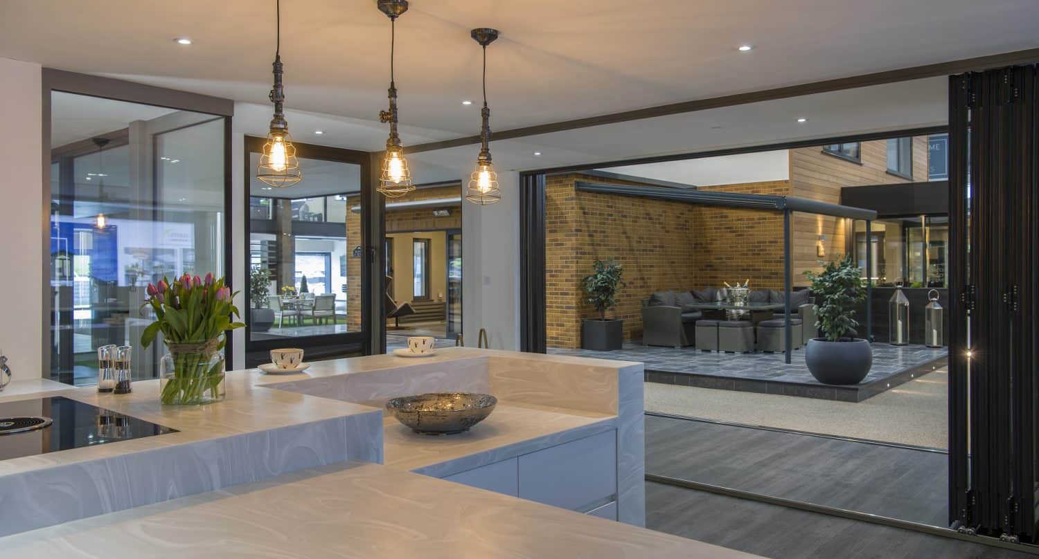 Modern open-plan kitchen with marble countertops, pendant lights, and a bowl centerpiece. The space opens to a stylish outdoor patio through sleek bifold doors, with large windows and brick walls creating an inviting indoor-outdoor living area.