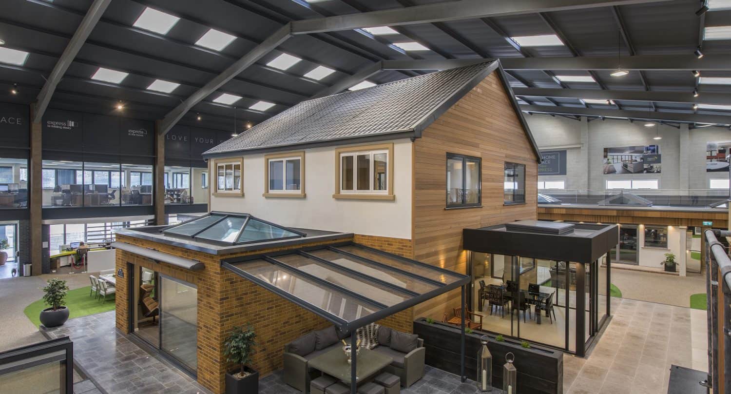 A modern showroom features a full-sized model house with large windows, bespoke glass solutions like a glass canopy, stylish front doors, and outdoor seating areas, all under a spacious, well-lit industrial ceiling.