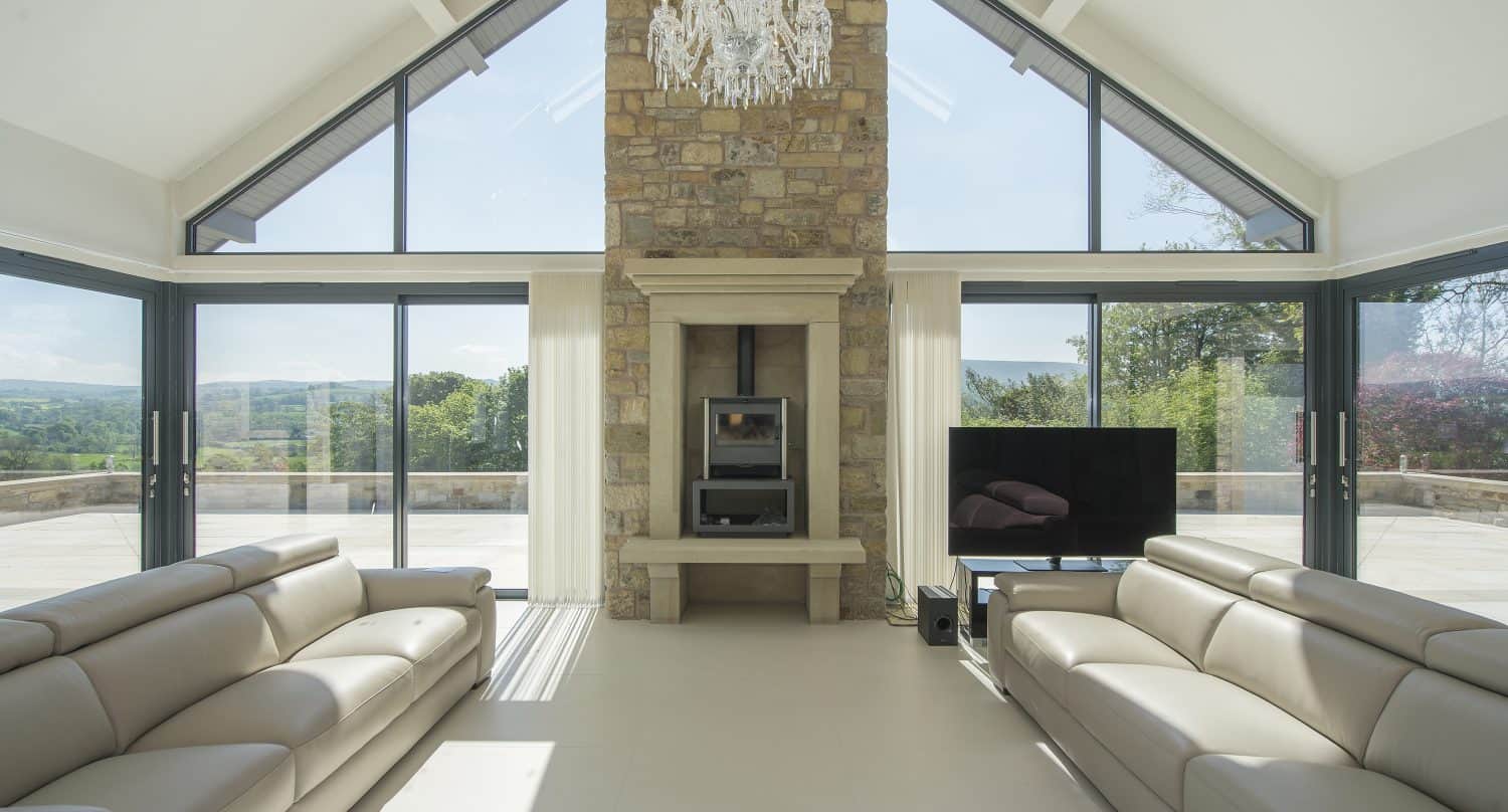 A spacious, modern living room with large sliding doors, beige leather sofas, a stone fireplace, a chandelier, and a flat-screen TV, overlooking a scenic outdoor view.