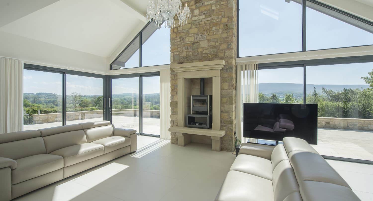 A bright, spacious living room with large windows, light-colored sofas, a stone fireplace, a chandelier, and stunning views of the countryside through floor-to-ceiling bifold doors.