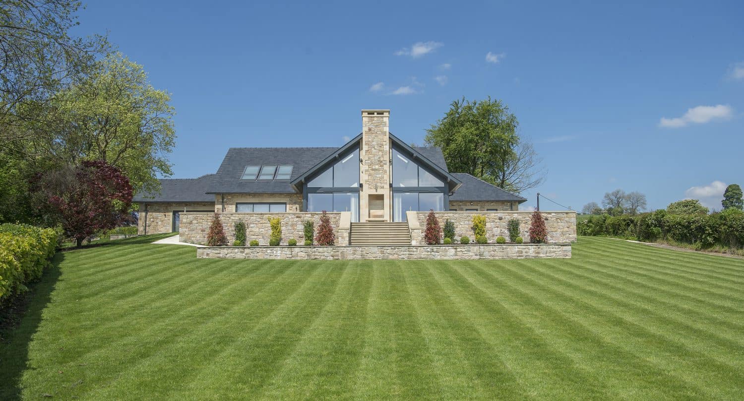Modern house with large glass windows, bifold doors, and a stone chimney, set on a lush, green, neatly mowed lawn, surrounded by trees and shrubs under a clear blue sky.