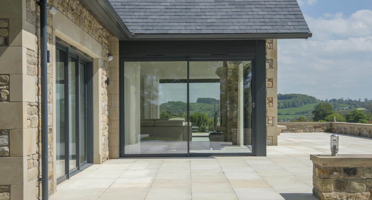 Modern house with large bifold doors, stone exterior, and spacious patio. Inside, a sofa is visible, and outside, scenic views of green hills and a partly cloudy blue sky complete this bespoke glass solution.