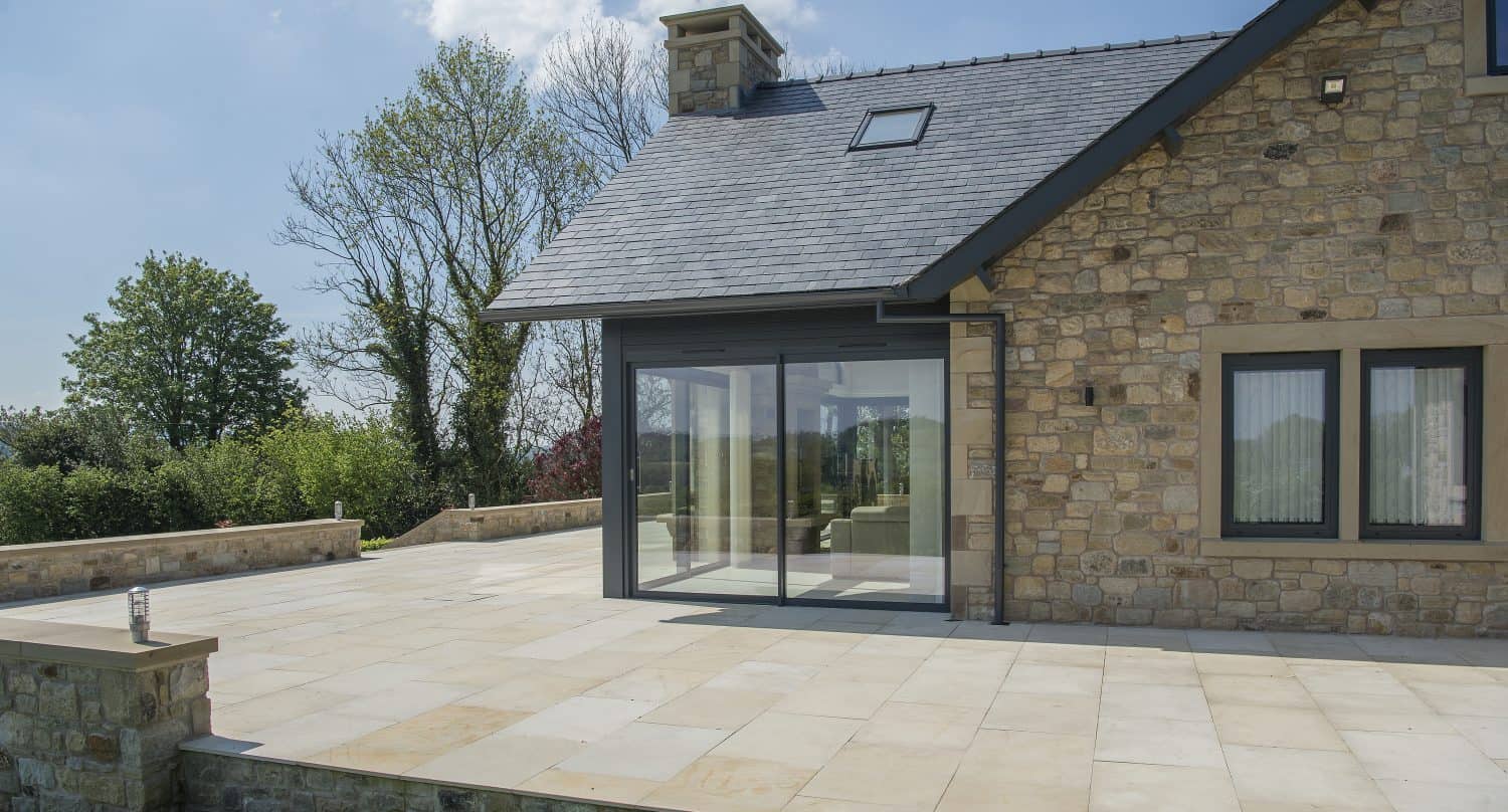 Modern stone house with bespoke glass solutions, featuring large sliding doors and windows, a spacious stone patio, and a well-maintained grassy lawn. Trees and a blue sky with scattered clouds complete the scenic backdrop.