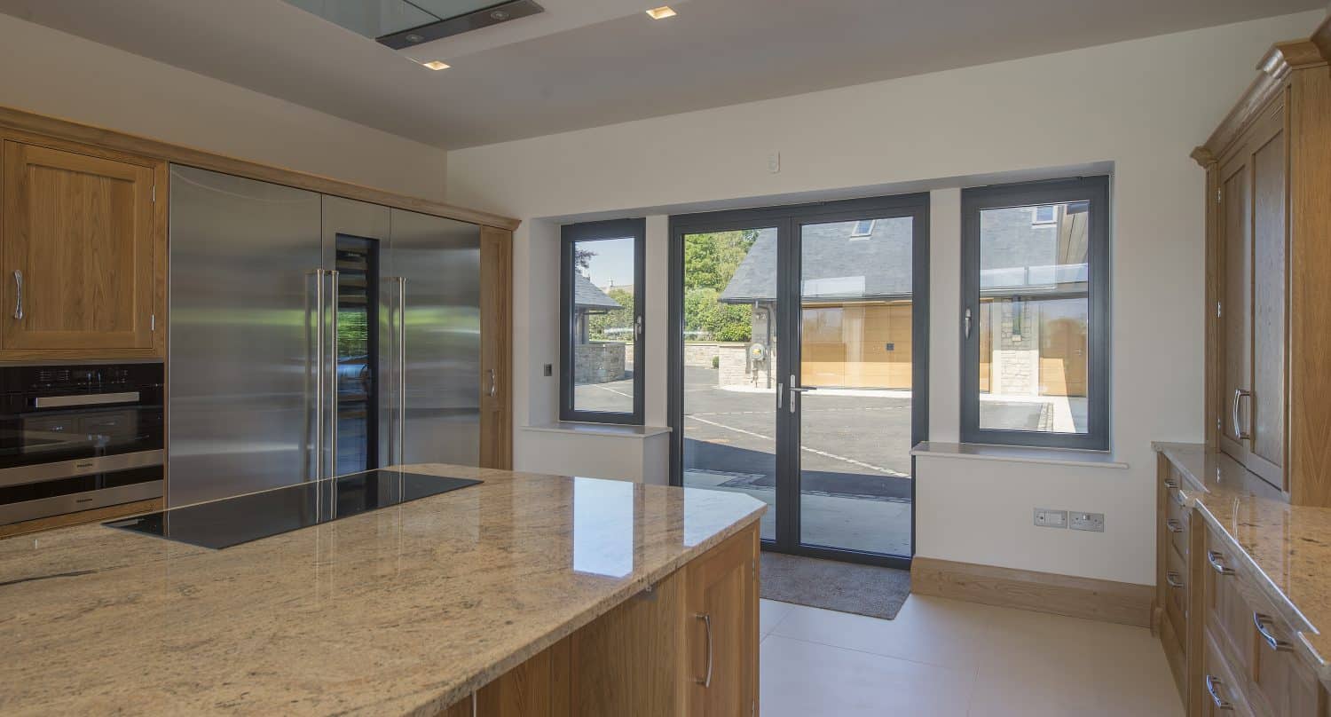 Modern kitchen with wooden cabinets, a large granite island countertop, stainless steel appliances, built-in oven, and expansive bespoke glass solutions including large windows and sliding doors overlooking buildings and surrounding greenery.