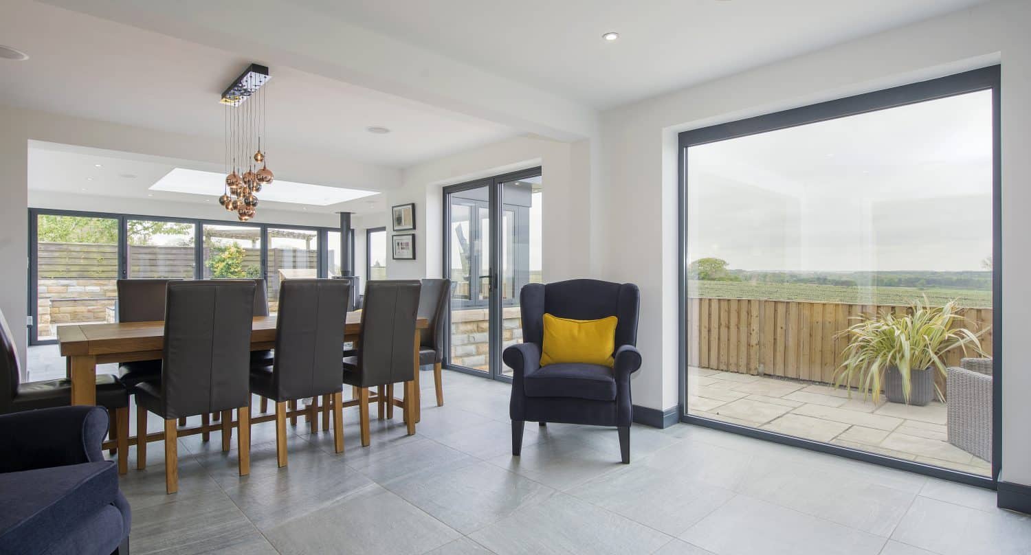 Modern dining area with a wooden table, black chairs, and a navy armchair with a yellow cushion. Large sliding doors open to a patio with outdoor seating and views of greenery beyond.